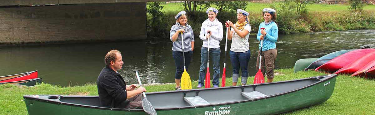 Einweisung. Kanu am Ufer, Einweiser sitzt mit Paddel im Boot, hinter dem Kanu 2 Frauen mit Paddeel am rechten Bildrand; dritte Frau am Rand halb verdeckt. Dahinter Neckar und Brückenpfeiler.